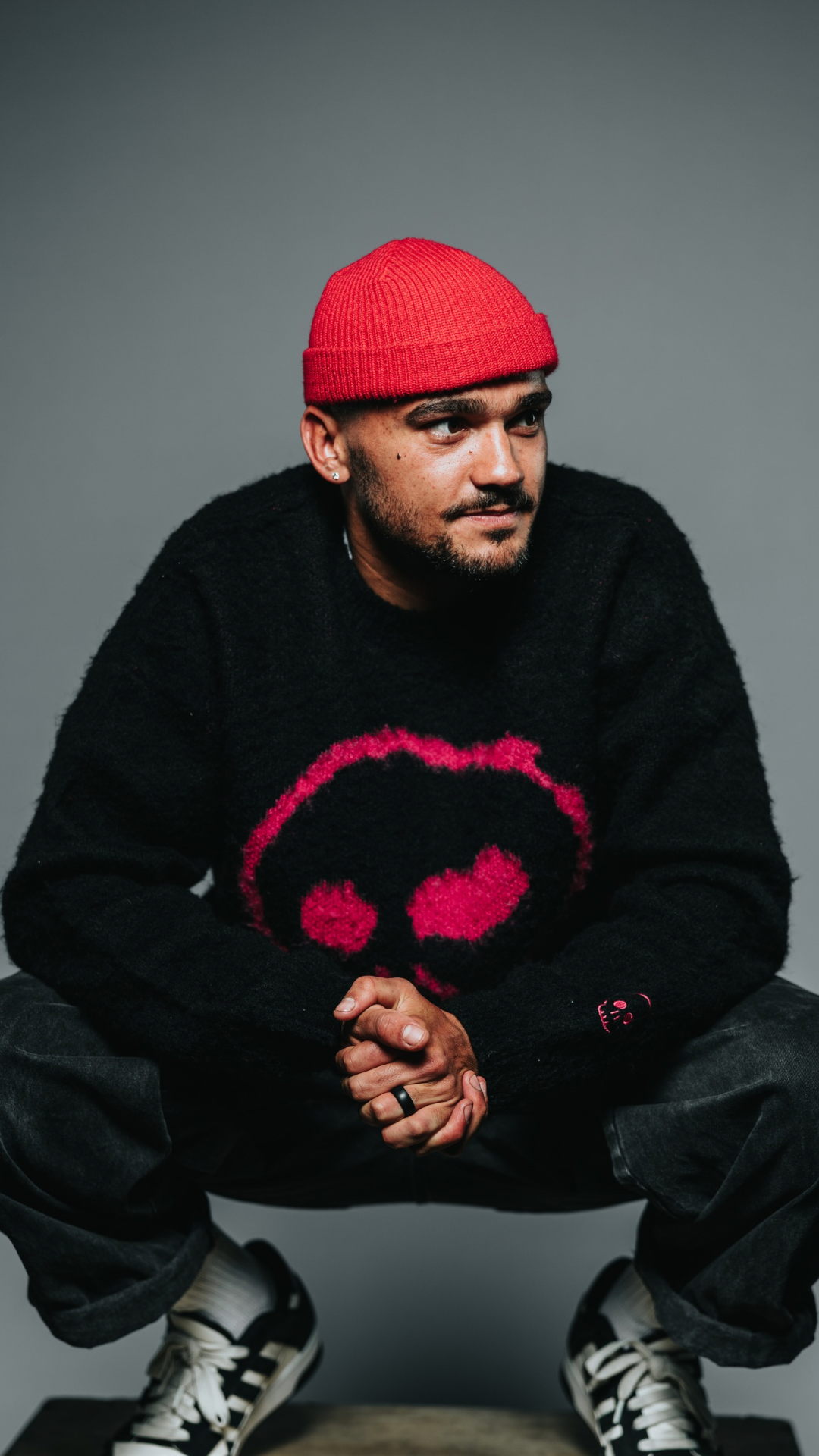 Man wearing a black sweater with a red logo and a red beanie sitting on a gray background