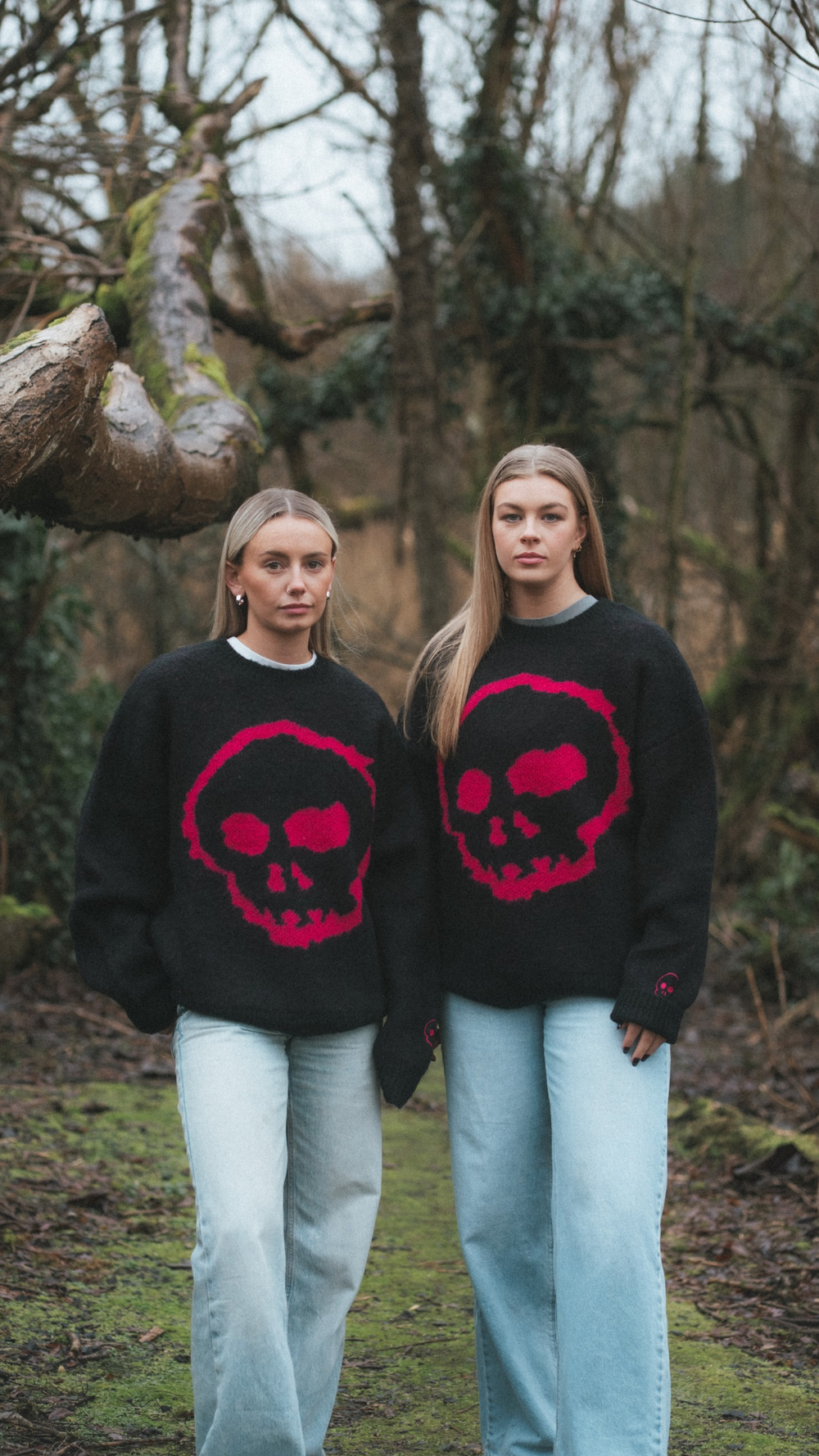 Two people wearing black sweaters with red skull designs in a forest setting.