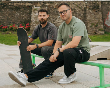 Two men sitting on a green bench with a skateboard, outdoors.