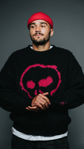 Man wearing a black sweater with a pink skull design and a red beanie against a gray background