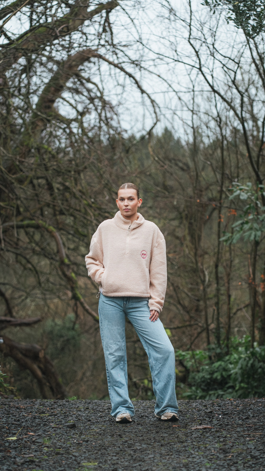 Person wearing a light pink sweater and blue jeans standing in a forest.