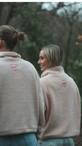 Two people wearing matching beige jackets with a pink logo in a forest setting.