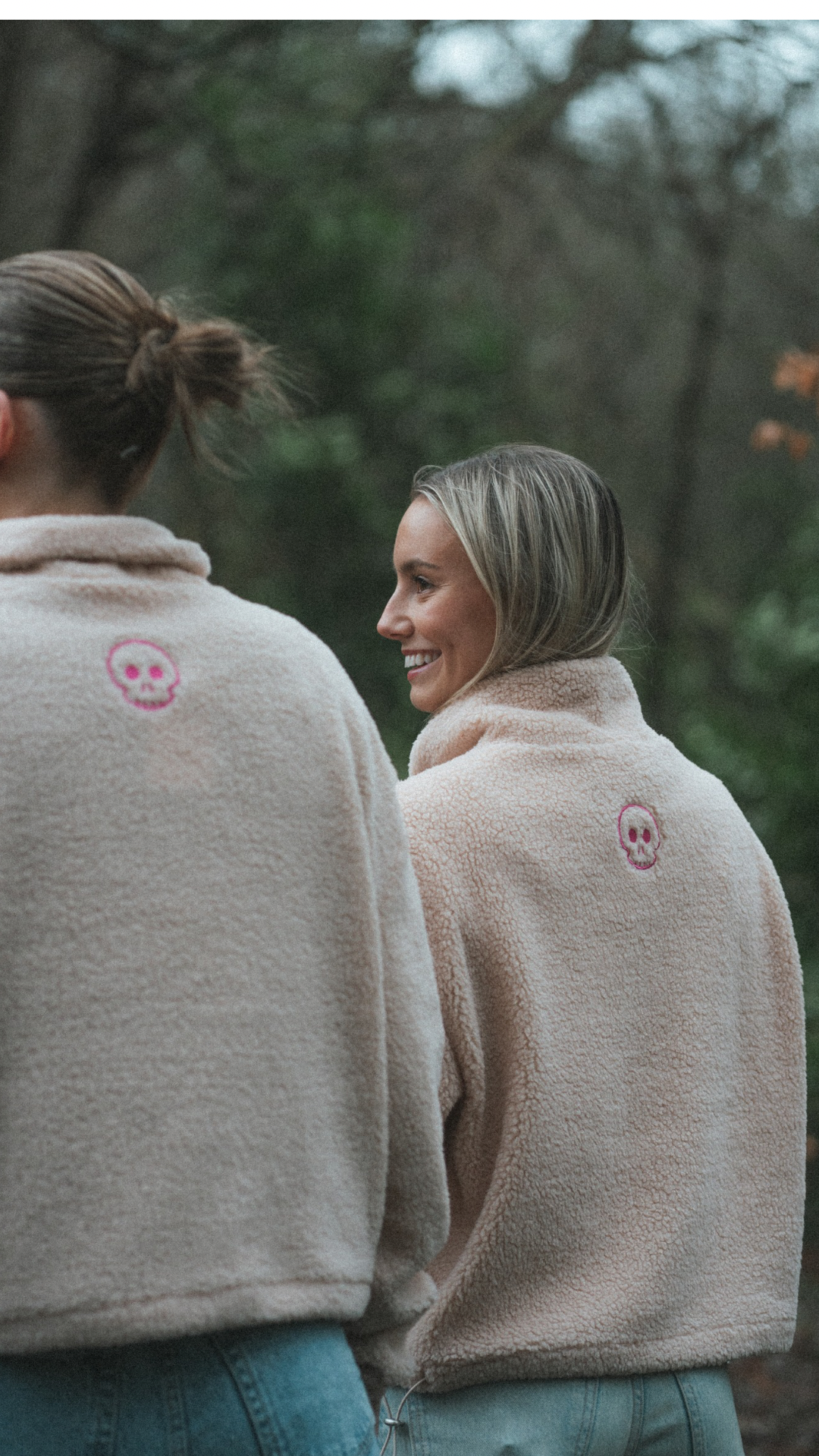 Two people wearing matching beige jackets with a pink logo in a forest setting.