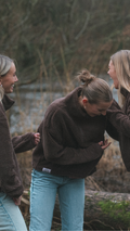 Three women standing outdoors by a body of water, laughing and interacting.