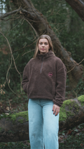 Person wearing a brown fleece jacket with a logo, standing in a forest setting.