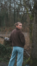 Person wearing a brown jacket and light blue jeans standing in a forest.