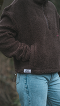 Brown fleece jacket with a brand label worn by a person, standing outdoors.