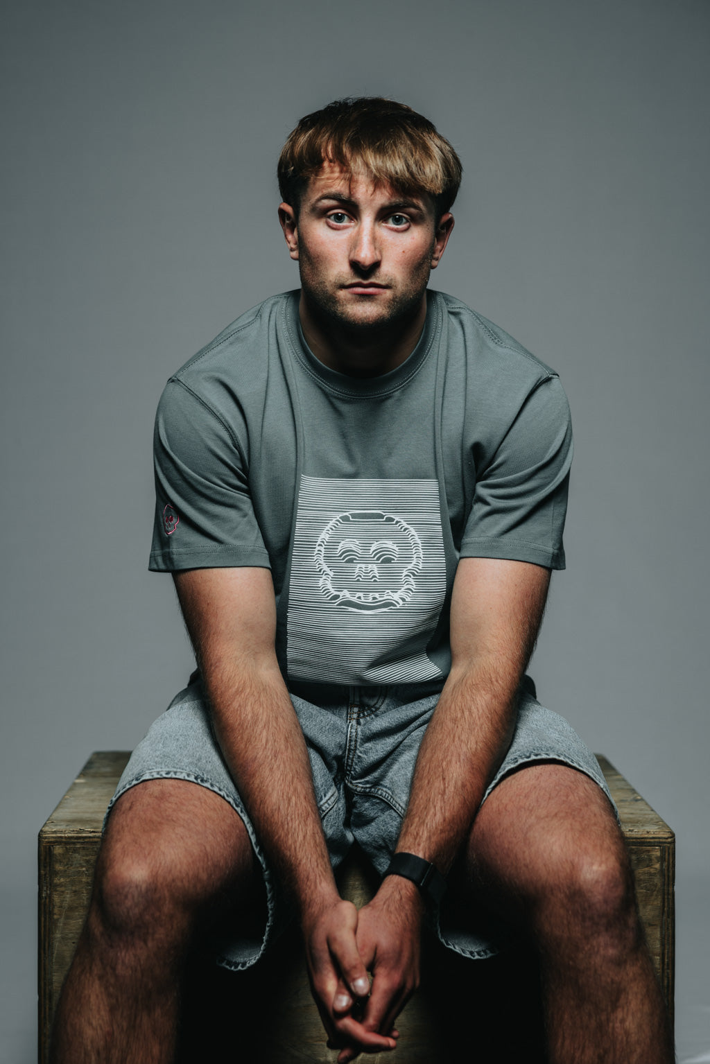 Man sitting on a wooden box wearing a gray t-shirt with a logo against a gray background